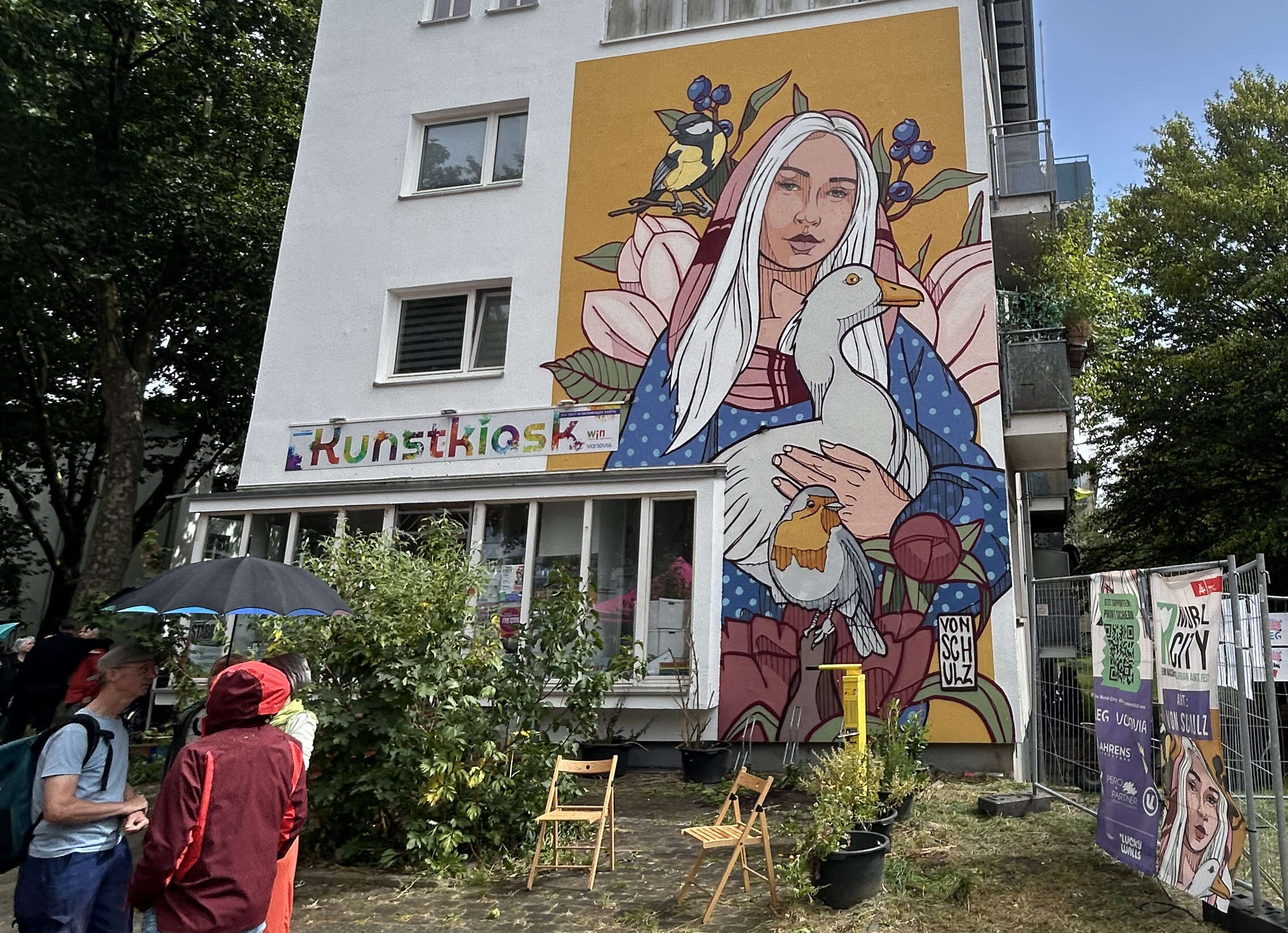Eine mit einem Fassadengemälde verzierte Hausfassade ist zu sehen. Darauf hält eine Frauengestalt eine Gans im Arm. Vor dem Gebäude befinden sich links ein paar Personen.