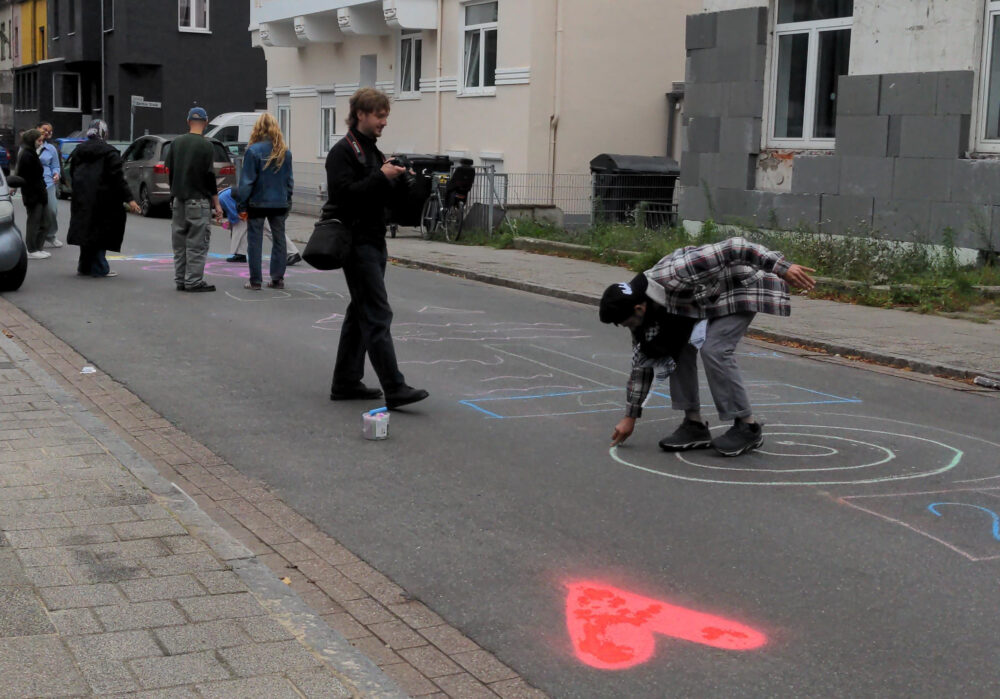 Mehrere Personen stehen auf einer Straße. Eine Person hält eine Kamera in der Hand, eine weitere Person besprüht die Straße mit Farben. Auf dem Straßenpflaster ist ein rotes Herz aufgesprüht.