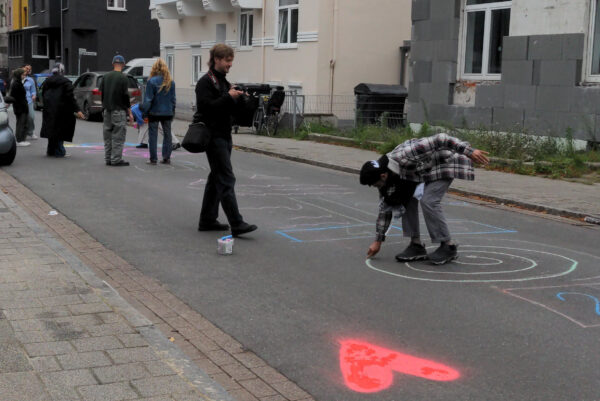 Mehrere Personen stehen auf einer Straße. Eine Person hält eine Kamera in der Hand, eine weitere Person besprüht die Straße mit Farben. Auf dem Straßenpflaster ist ein rotes Herz aufgesprüht.