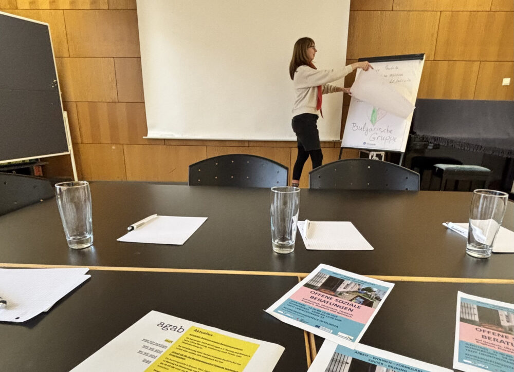 Im Hintergrund befindet sich eine Person vor einer Leinwand und schlägt an einem Flipchart ein Blatt um. Zur linken steht eine Pinwand. Im Vordergrund befinden sich ein Tischblock mit Flyern und Wassergläsern.