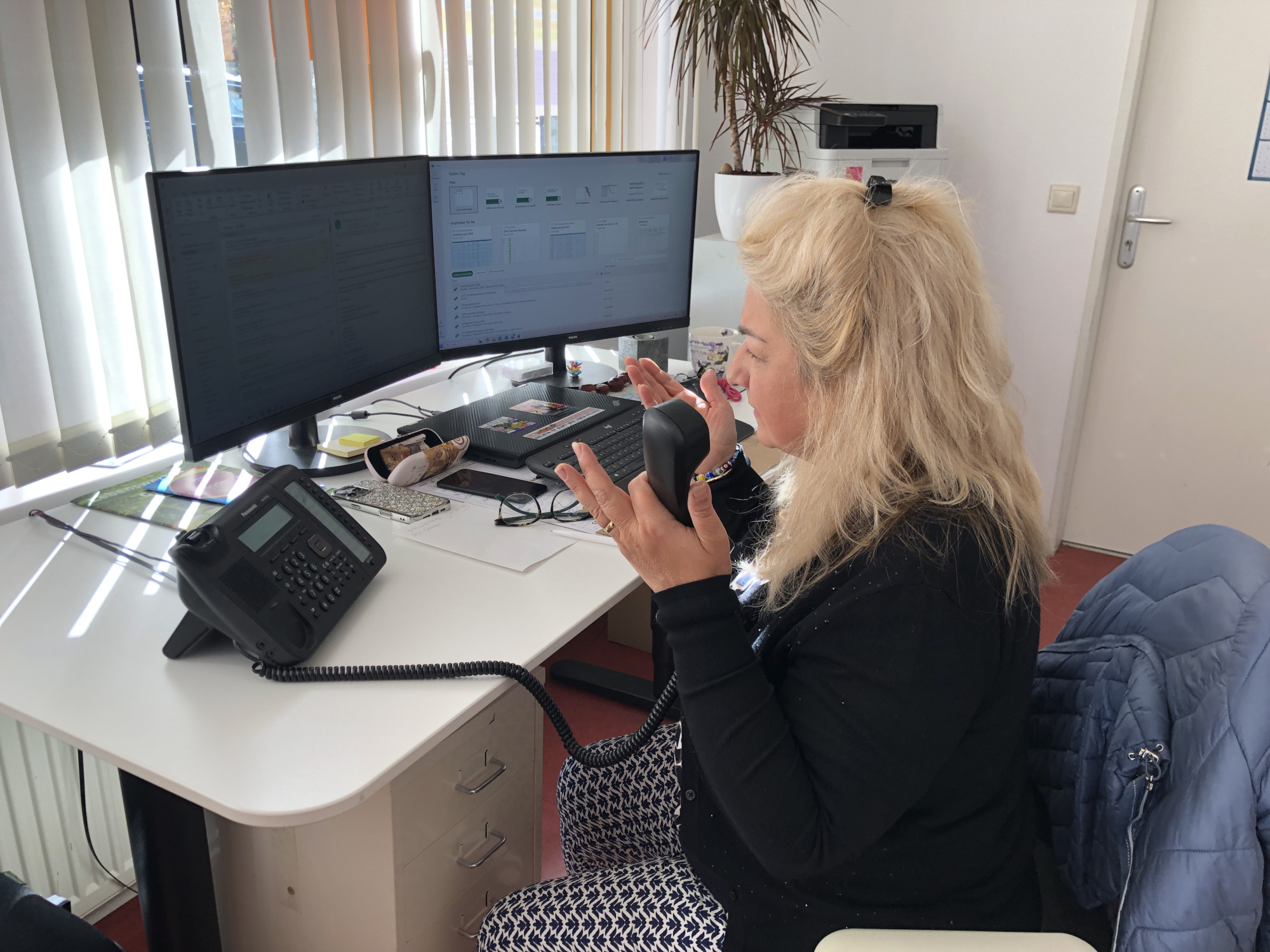 Eine Person sitzt an einem Schreibtisch mit zwei Bildschirmen und hält den telefonhörer in der Hand. Mit der anderen Hand macht sie eine hilflose Geste der Verzweiflung.