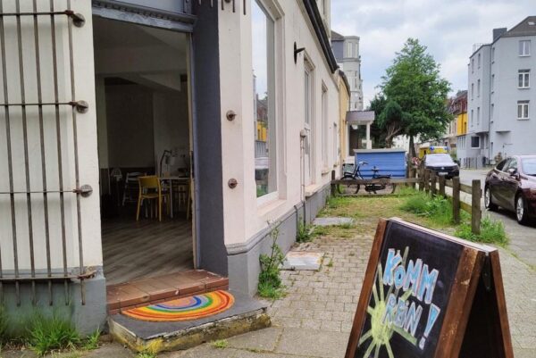 Die Tür zu einem Ladenlokal steht weit offen. Auf der Türschwelle liegt eine bunte Fußmatte. Vor dem Lokal steht eine Klapptafel auf der steht "Herzlich Wilkommen".