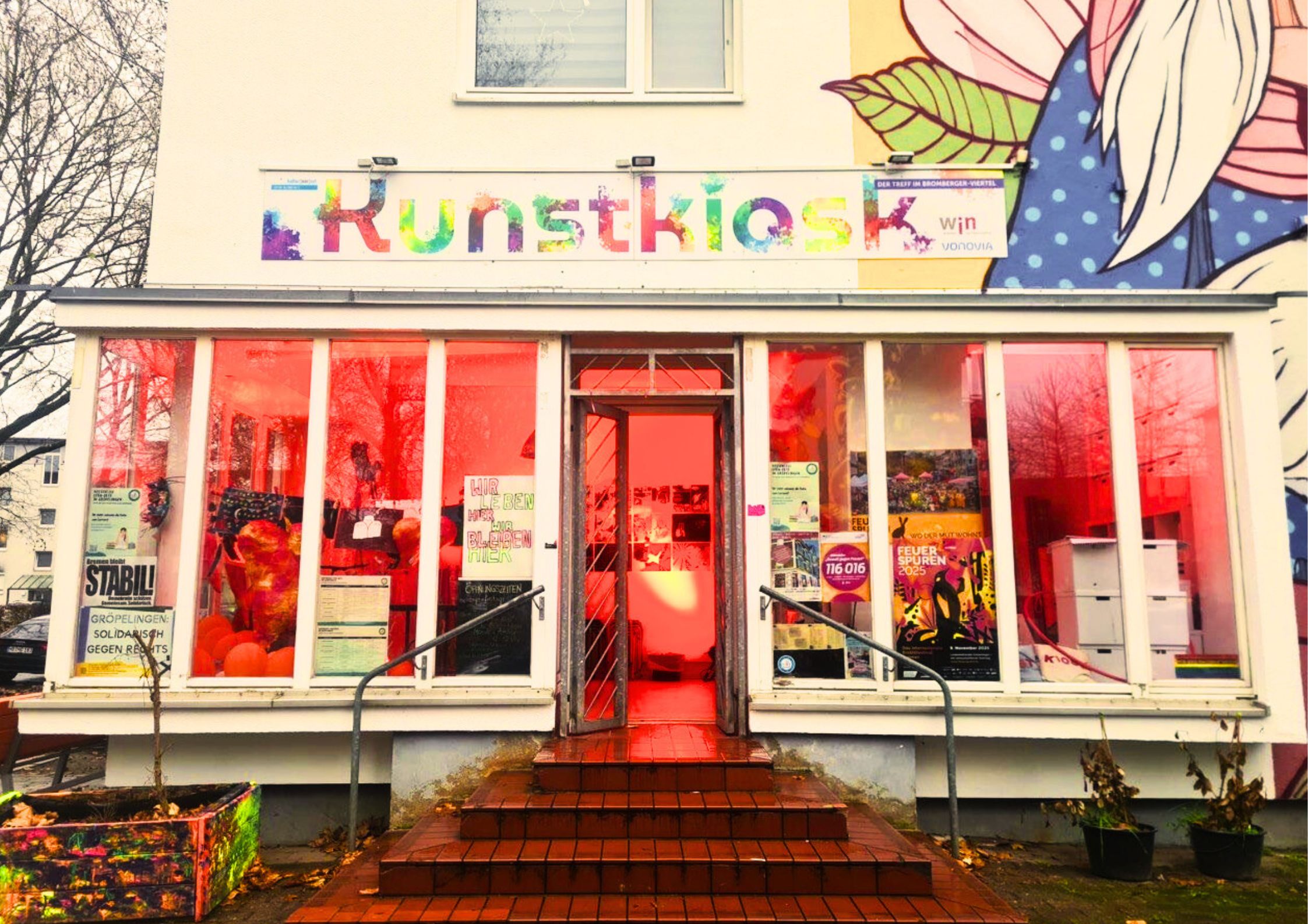 Ein Ladenlokal leuchtet orange durch die ausladenden Fensterfronten. Über dem Ladenlokal steht der Schriftzug "Kunstkiosk".