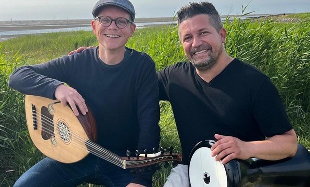 Zwei Männer mit Instrumenten lachen in die Kamera. Der eine hat ein Oud der andere spielt eine Trommel.