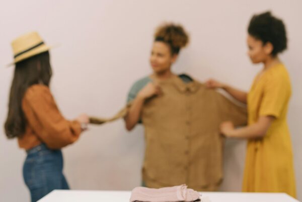 Vorne auf einem Tisch liegen ordentlich gefaltete Stoffe, ein Laptop und ein kleines Nähset. Im Hintergrund halten drei Frauen gemeinsam ein braunes Kleidungsstück hoch.