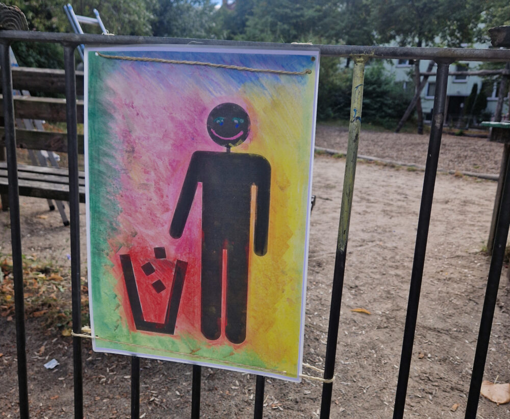 Auf einem Schild an einem Zaun ist ein Pikrogramm von einem Menschen, der Müll in einen Mülleimer entsorgt.