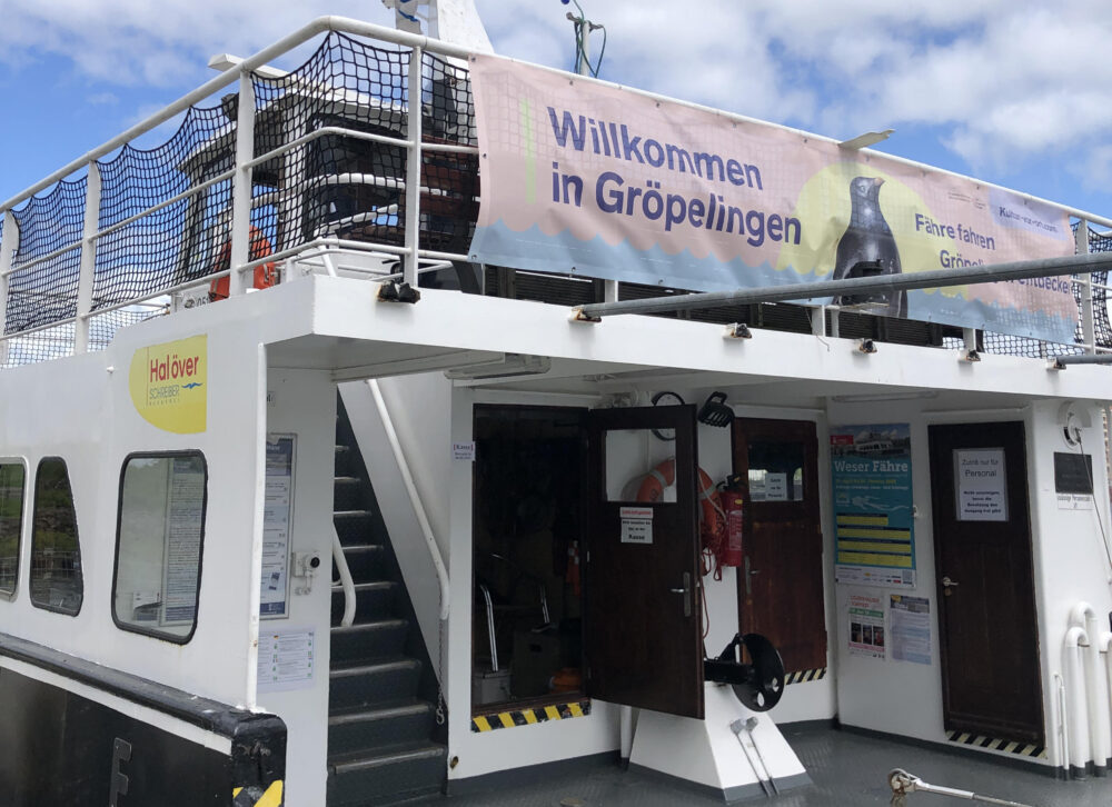 Ein Blick auf das Fahrgastschiff Pusdorp, das als Weserfähre Gröpelingen, Überseestadt und WQoltmershausen verbindet. Ein Banner mit einem Pinguin und dem Spruch "Willkommen in Gröpelingen" ist an der Reeling befestigt.
