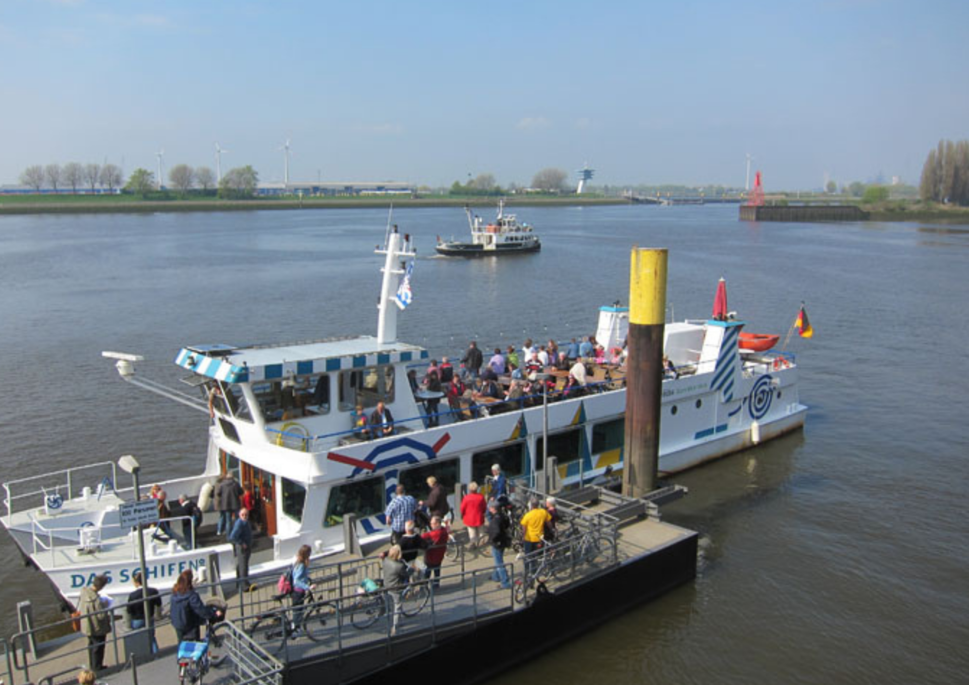 Ein Fahrgastschiff am Anleger. Fahrgäste steigen ein und aus. Im hintergrund ist ein weiteres Schiff zu sehen und der markante Radarturm am Lankenauer Höft