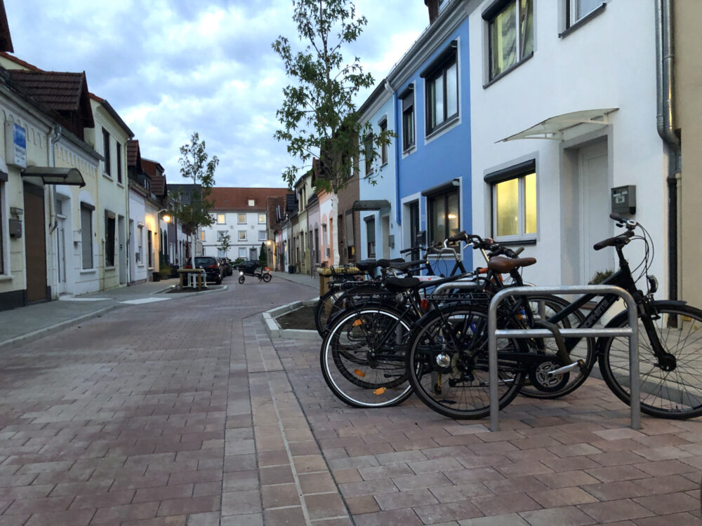 Zu sehen ist die Gestaltung der Rasteder Straße in Bremen Gröpelingen nach der Klimaangepassten Sanierung: ein lockeres Plaster, neue Bäume, Fahrradparkplatze und Parkbuchten sind zu sehen.