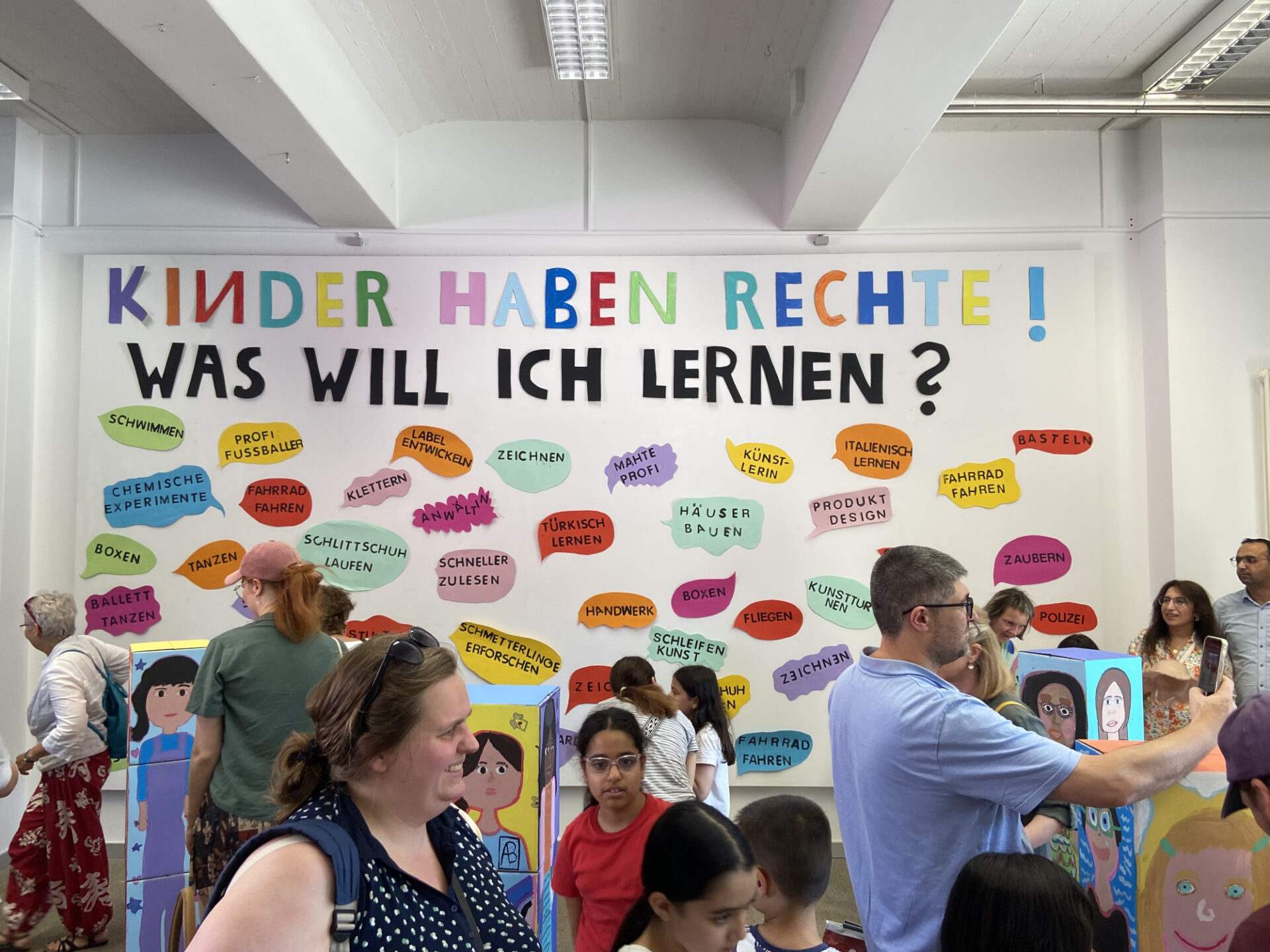 In einer Galerie sind viele Menschen, darunter auch viele Kinder. An einer großen weißen Wand steht der Satz: "Kinder haben Rechte! Was will ich lernen?" Darunter stehen Sprechblasen auf denen ganz unterschiedliche Lernwünsche stehen wie zum Beispiel Schwimmen oder Italienisch lernen.