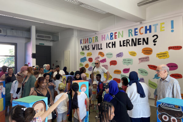 In einer Galerie sind viele Menschen, darunter auch viele Kinder. An einer großen weißen Wand steht der Satz: "Kinder haben Rechte! Was will ich lernen?" Darunter stehen Sprechblasen auf denen ganz unterschiedliche Lernwünsche stehen wie zum Beispiel Schwimmen oder Italienisch lernen.