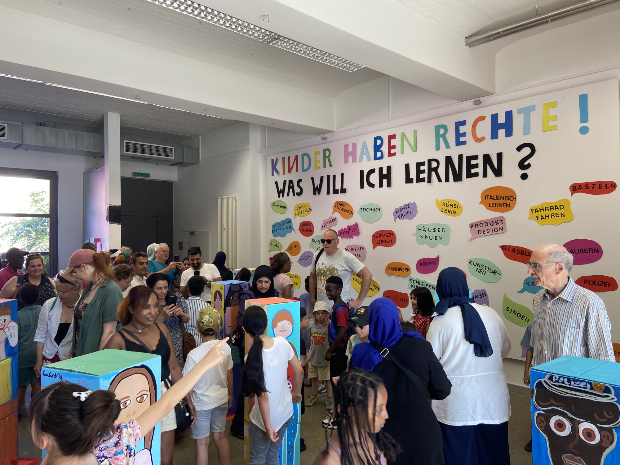 In einer Galerie sind viele Menschen, darunter auch viele Kinder. An einer großen weißen Wand steht der Satz: "Kinder haben Rechte! Was will ich lernen?" Darunter stehen Sprechblasen auf denen ganz unterschiedliche Lernwünsche stehen wie zum Beispiel Schwimmen oder Italienisch lernen.