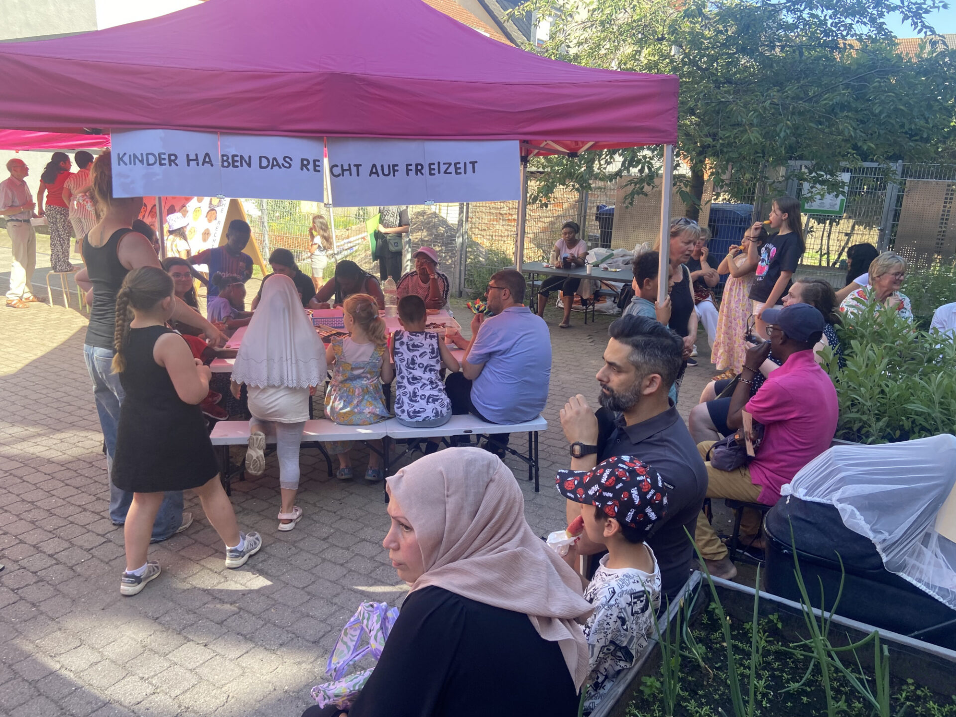 Auf einem gepflasterten Hof befindet sich ein pinker Pavillon, an dem eine Mitmachaktion stattfindet. In großen Buchstaben steht auf einem Spruchband: "Kinder haben das Recht auf Freizeit". Viele Personen, darunter viele Kinder beteiligen sich. Weitere Personen sitzen gemütlich auf Bänden und schauen zu.