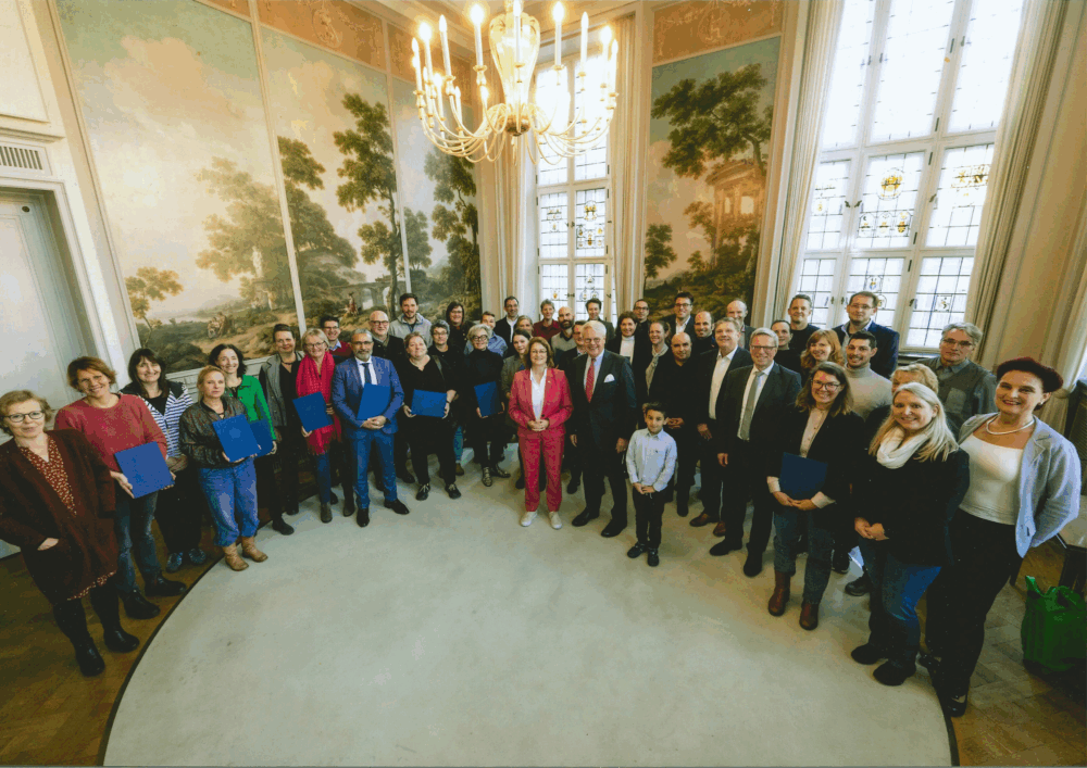 Eine große Gruppe von Personen steht in einem festlich gestalteten Raum mit hohen Fenstern und Wandgemälden. Im Zentrum des Bildes befinden sich mehrere Personen in formeller Kleidung, einige halten blaue Mappen in den Händen. Ein Kronleuchter hängt von der Decke, das Licht ist hell und die Stimmung wirkt feierlich. Die Gruppe ist halbkreisförmig aufgestellt und blickt geschlossen in die Kamera.