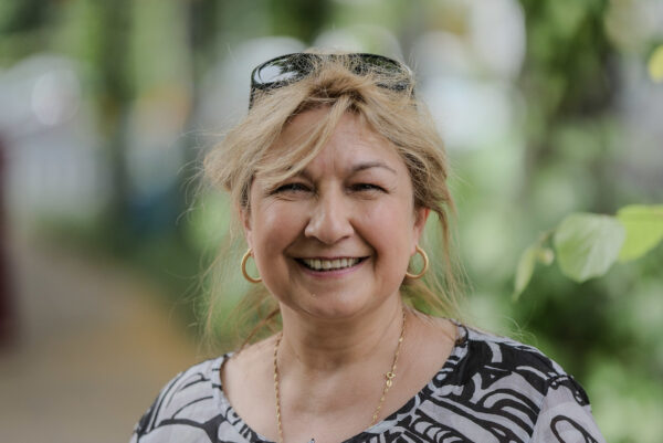 Eine Person in bunt gemusterten Oberteil, lässig ins Haar hochgeschobener Sonnenbrille lächelt in die Kamera.