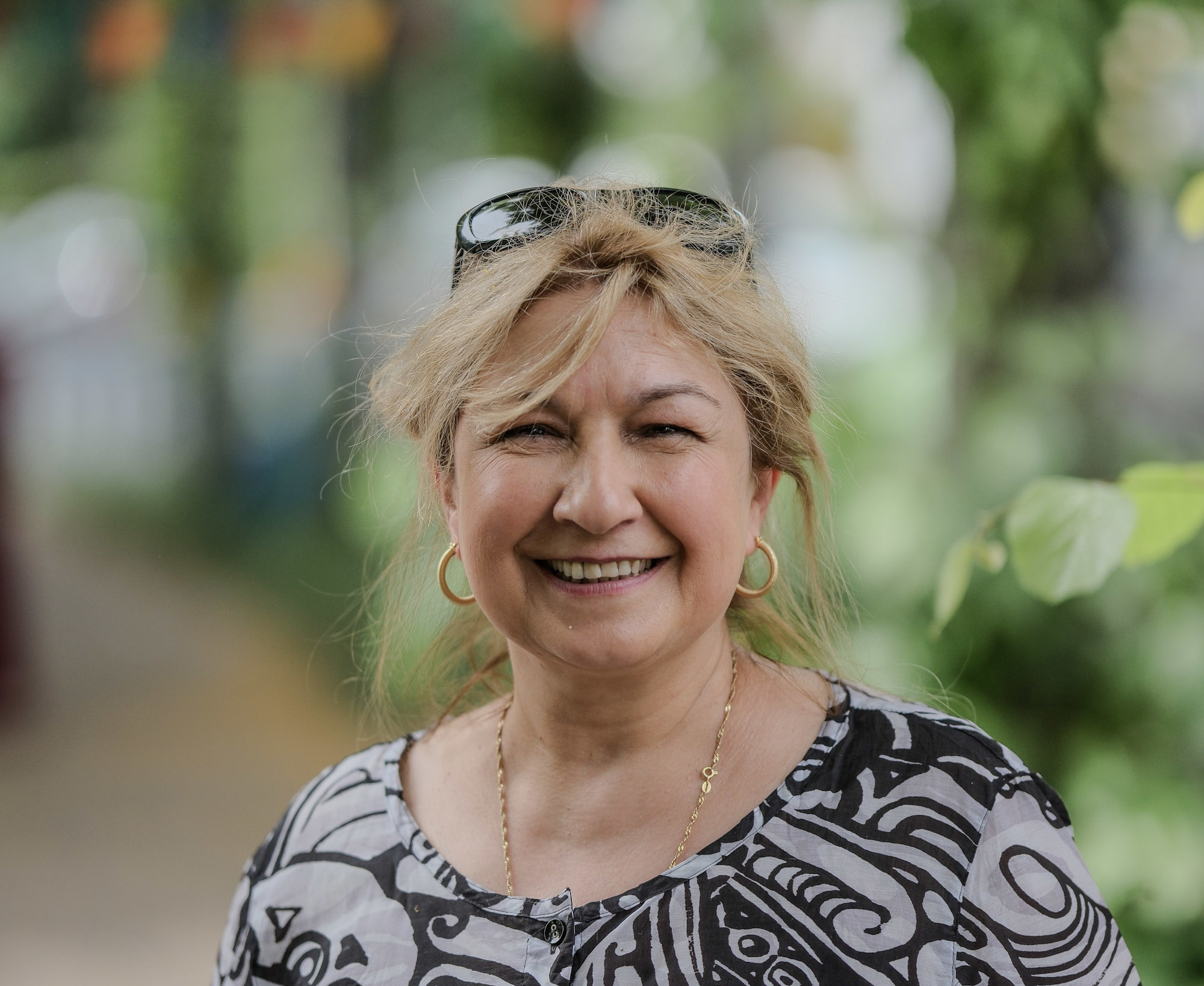Eine Person in bunt gemusterten Oberteil, lässig ins Haar hochgeschobener Sonnenbrille lächelt in die Kamera.