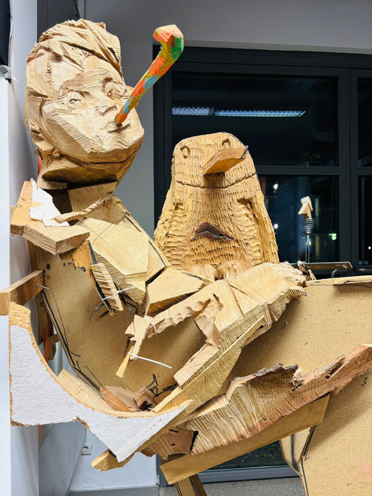 Zwei Skulpturen aus Sperrholz und anderem Holz sind hier in Nahaufnahme zu sehen. Die eine stellt eine Person dar mit einer Tröte im Mund, die andere einen Pinguin.