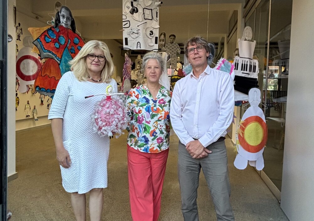 Vor einem Ausstellungsraum stehen drei Personen und lächeln in die Kamera. Im hintergrund sind Ausstellugnsstücke zu sehen. Die Person in der Mitte hält einen Blumenstraß in der Hand.