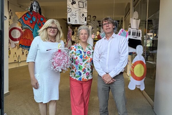 Vor einem Ausstellungsraum stehen drei Personen und lächeln in die Kamera. Im hintergrund sind Ausstellugnsstücke zu sehen. Die Person in der Mitte hält einen Blumenstraß in der Hand.
