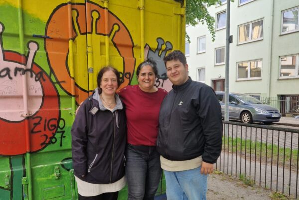 Vor einem bunt angemalten Container stehen drei Personen dicht nebeneinander und lächeln in die Kamera.