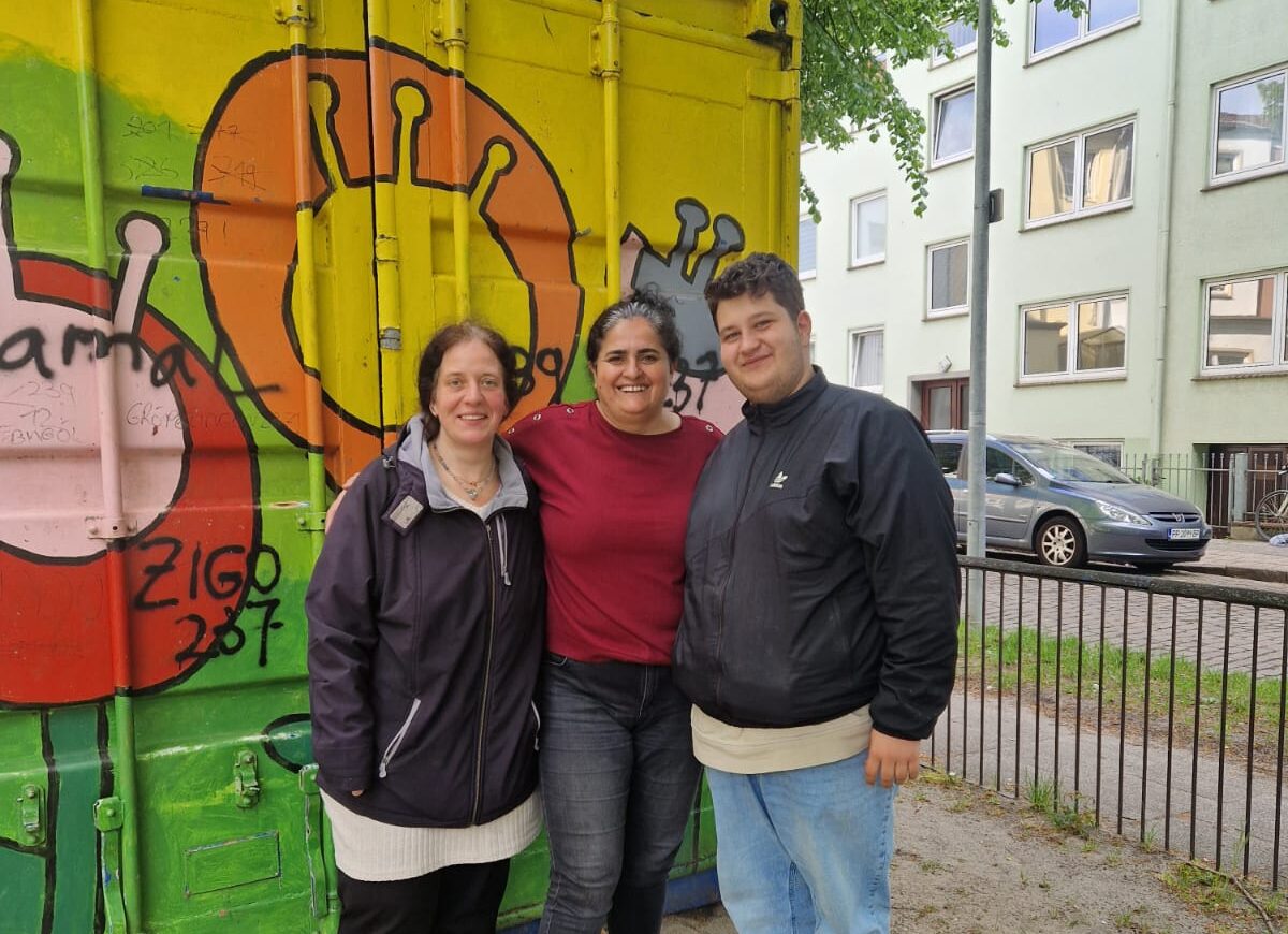 Vor einem bunt angemalten Container stehen drei Personen dicht nebeneinander und lächeln in die Kamera.