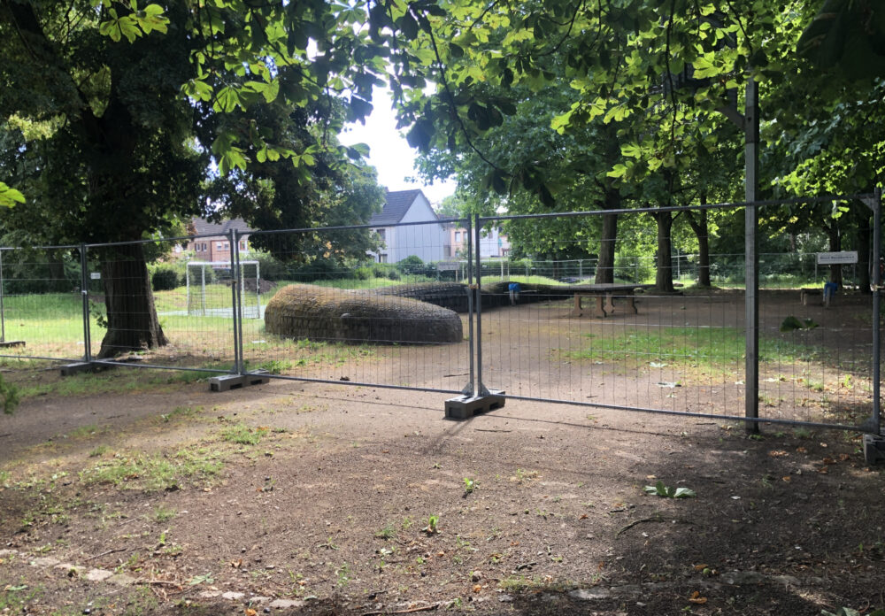 Zwischen Bäumen ist ein Spielplatz zu erkennen, der durch Bauzäune abgesperrt ist.