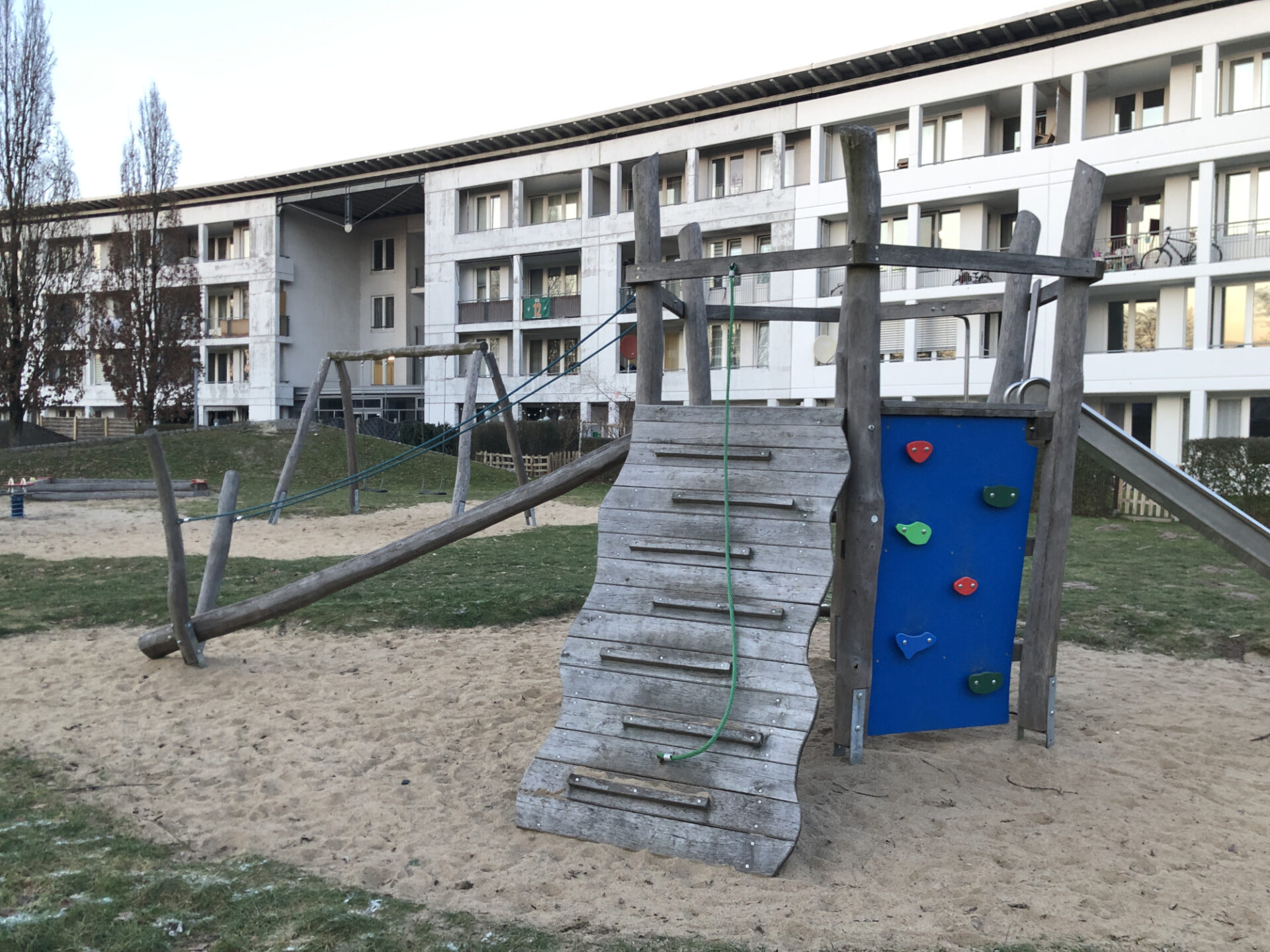 Vor einem großen Mehrparteienhaus befindet sich ein Spielplatz mit Rutsche, Kletterwand, schaukeln und Wippen.