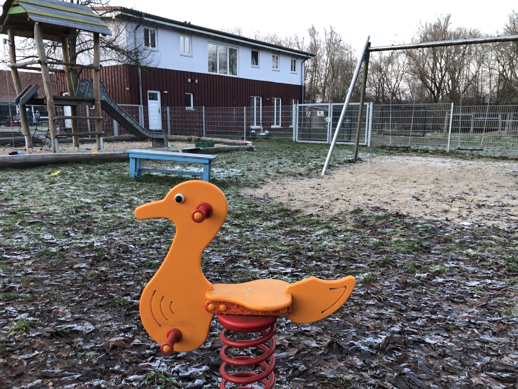 Das Bild zeigt einen Spielplatz. Im Vordergrund befindet sich eine Wippe für Kleinkinder in Form einer orangenen Ente. Links ist ein Haus mit Rutsche, rechts befindet sich eine Schaukel. Im Hintergrund sind ein Gebäude und Bäume zu sehen.