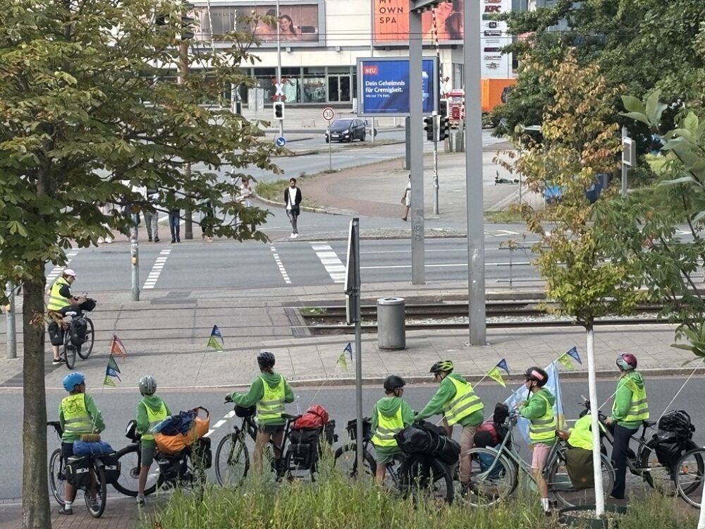 Zwischen zwei Bäumen mit grünen Baumkronen warten Fahrradfahrende gemeinsam vor einer Ampel an einer mehrspurigen Straße. Alle tragen gelbe Warnwesten und Fahrradhelme.