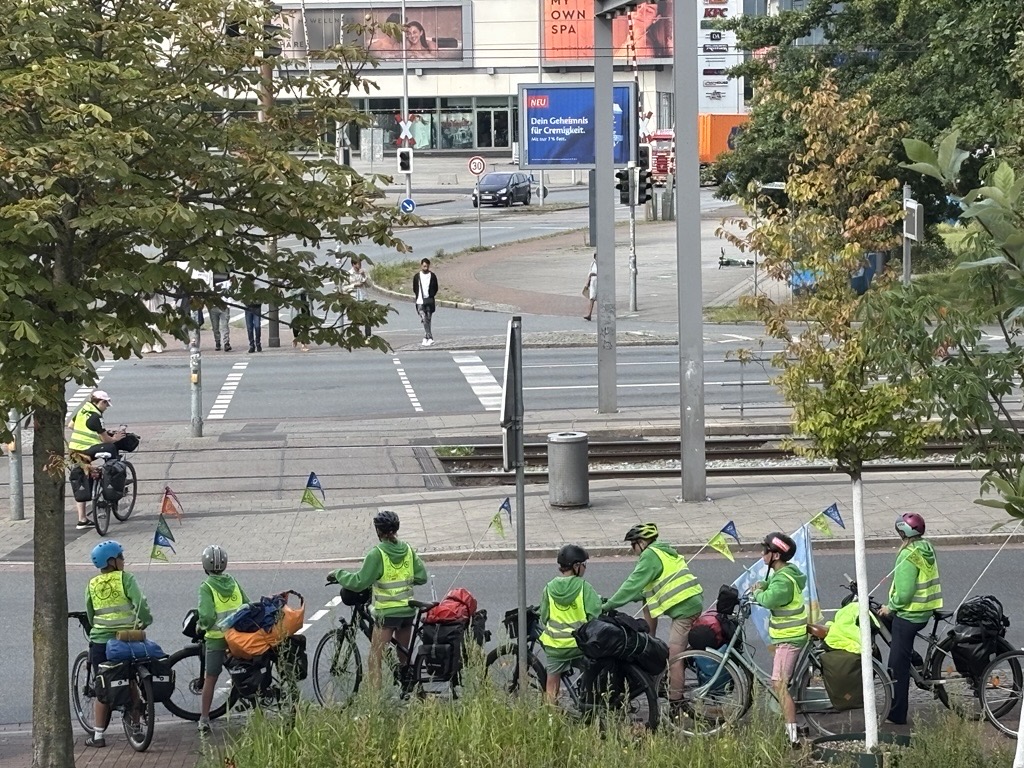 Zwischen zwei Bäumen mit grünen Baumkronen warten Fahrradfahrende gemeinsam vor einer Ampel an einer mehrspurigen Straße. Alle tragen gelbe Warnwesten und Fahrradhelme.