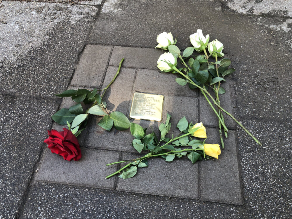 Ein glänzender Stolperstein ist von Blumen umrahmt.