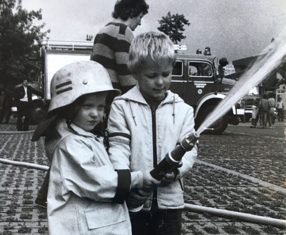 Zwei Kinder halten zusammen einen Feuerwehrschlauch, aus dem Wasser spritzt.