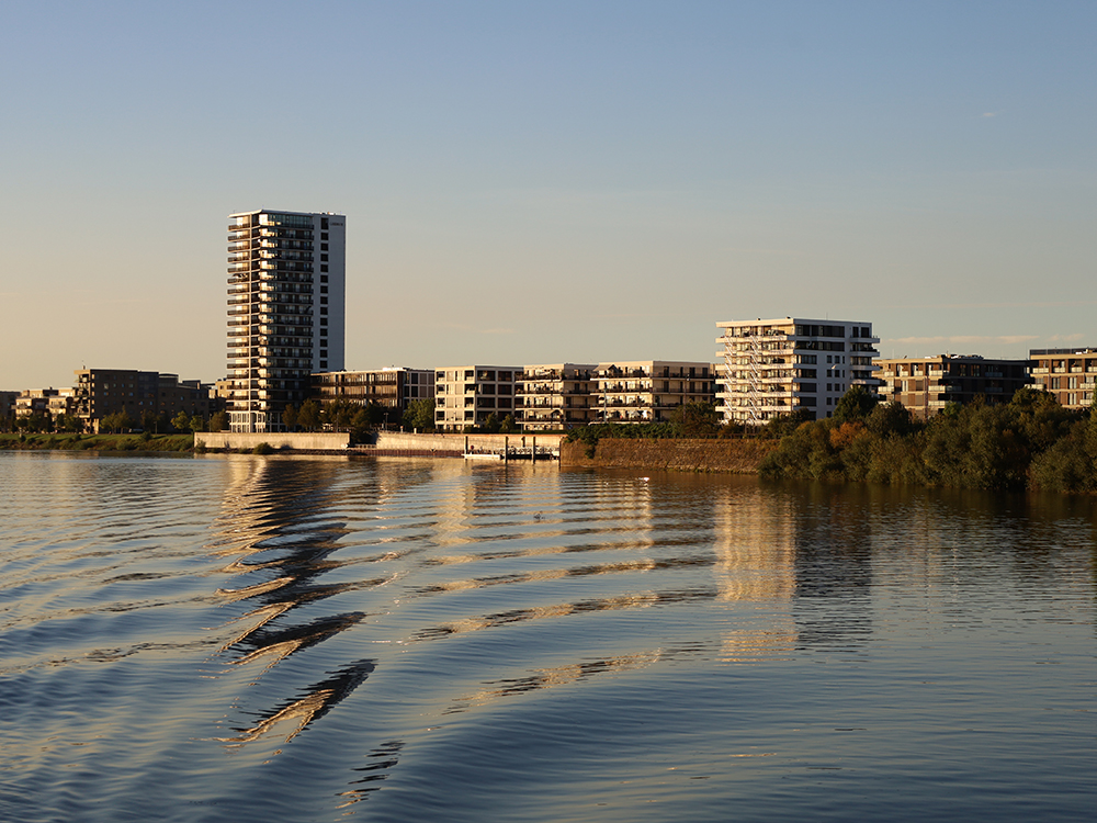 Abendstimmung von der Flusseite aus mit Blick auf ein Ufer mit Hochhäusern und Wohnhäusern.