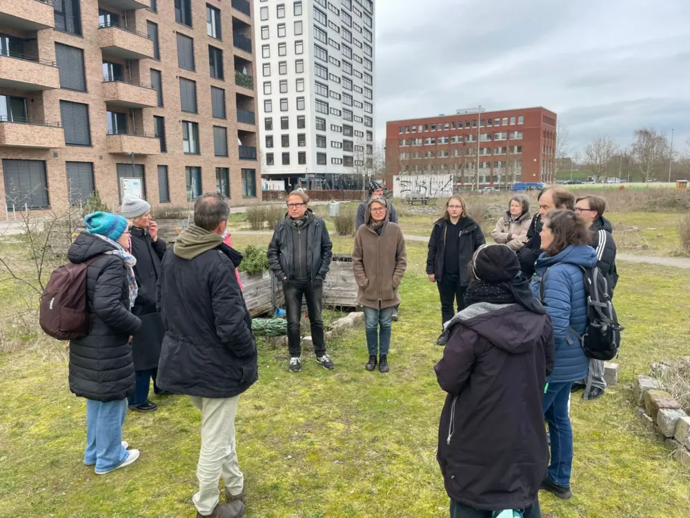 Eine Gruppe von warm gekleideten Personen steht im Halbkreis auf einer Wiese. Im Hintergrund ind Hochhäuser zu sehen.