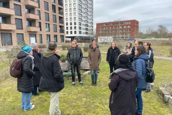 Eine Gruppe von warm gekleideten Personen steht im Halbkreis auf einer Wiese. Im Hintergrund ind Hochhäuser zu sehen.