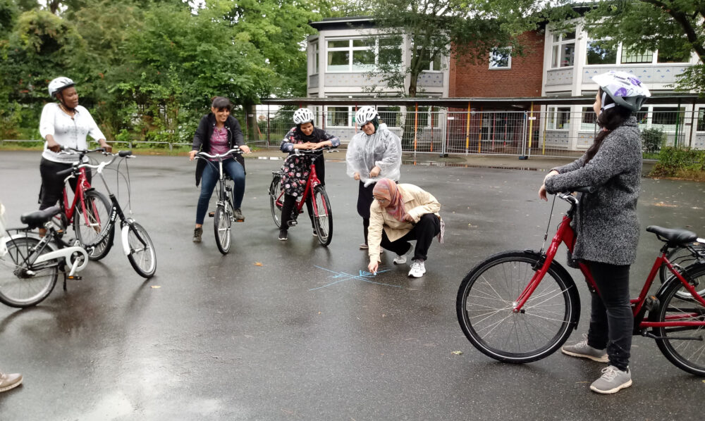 Auf einem Schulhof befinden sich mehrere Frauen mit Fahrradhelmen und Fahrrädern. Sie schauen einer weiteren Frau zu, die etwas auf den Asphalt mit Kreide zeichnet. Im Hintergrund befinden sich ein Gebäude und Büsche.