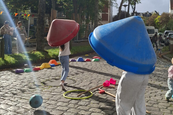 Auf dem Kopfsteinplaster einer Straße spielen verschieden Kinder. Zwei davon haben sich Objekte über den Kopf gestülpt, im Hintergrund wird Fahrrad gefahren. Am Rad stehen zwei Erwachsene.