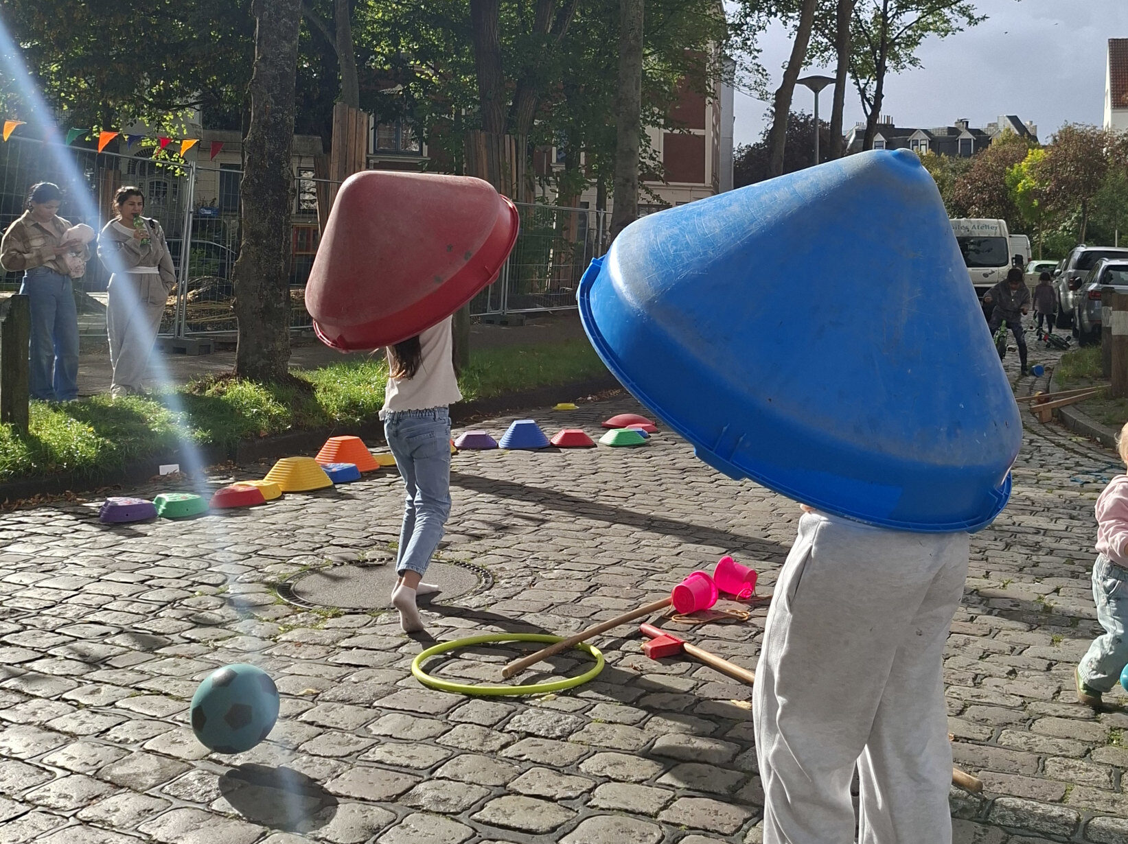 Auf dem Kopfsteinplaster einer Straße spielen verschieden Kinder. Zwei davon haben sich Objekte über den Kopf gestülpt, im Hintergrund wird Fahrrad gefahren. Am Rad stehen zwei Erwachsene.