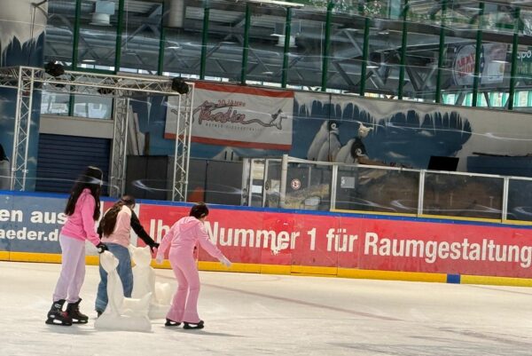 Drei weiblich zu lesende junge Personen sind beim Schlittschuhlaufen in einer großen Schlittschuhhalle.