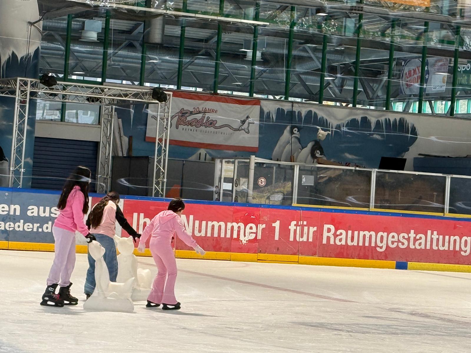 Drei weiblich zu lesende junge Personen sind beim Schlittschuhlaufen in einer großen Schlittschuhhalle.