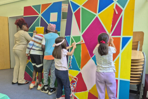 Fünf Kinder und Jugendliche stehen vor einer großen Platte, die sie Mosaikförmig farbig anmalen.