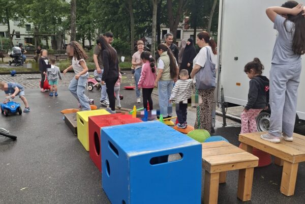 Auf der Straße steht rechts ein weißer Hänger. Davor befinden sich farbige Kisten und kleine Hocker. Rundherum befinden sich mehrere Kinder und Erwachsene.