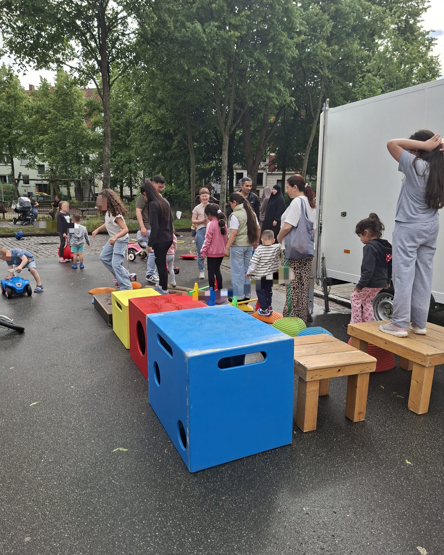 Auf der Straße steht rechts ein weißer Hänger. Davor befinden sich farbige Kisten und kleine Hocker. Rundherum befinden sich mehrere Kinder und Erwachsene.