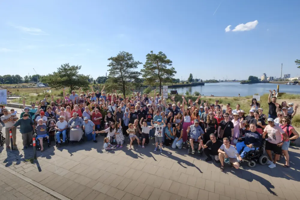 Das ist ein Gruppenfoto von allen Beiligten beim Sommerfest in der Überseestadt "Zu Tisch" Die Personen winken und lächeln in die Kamera. Im Hintergrund befindet sich ein Fluss.