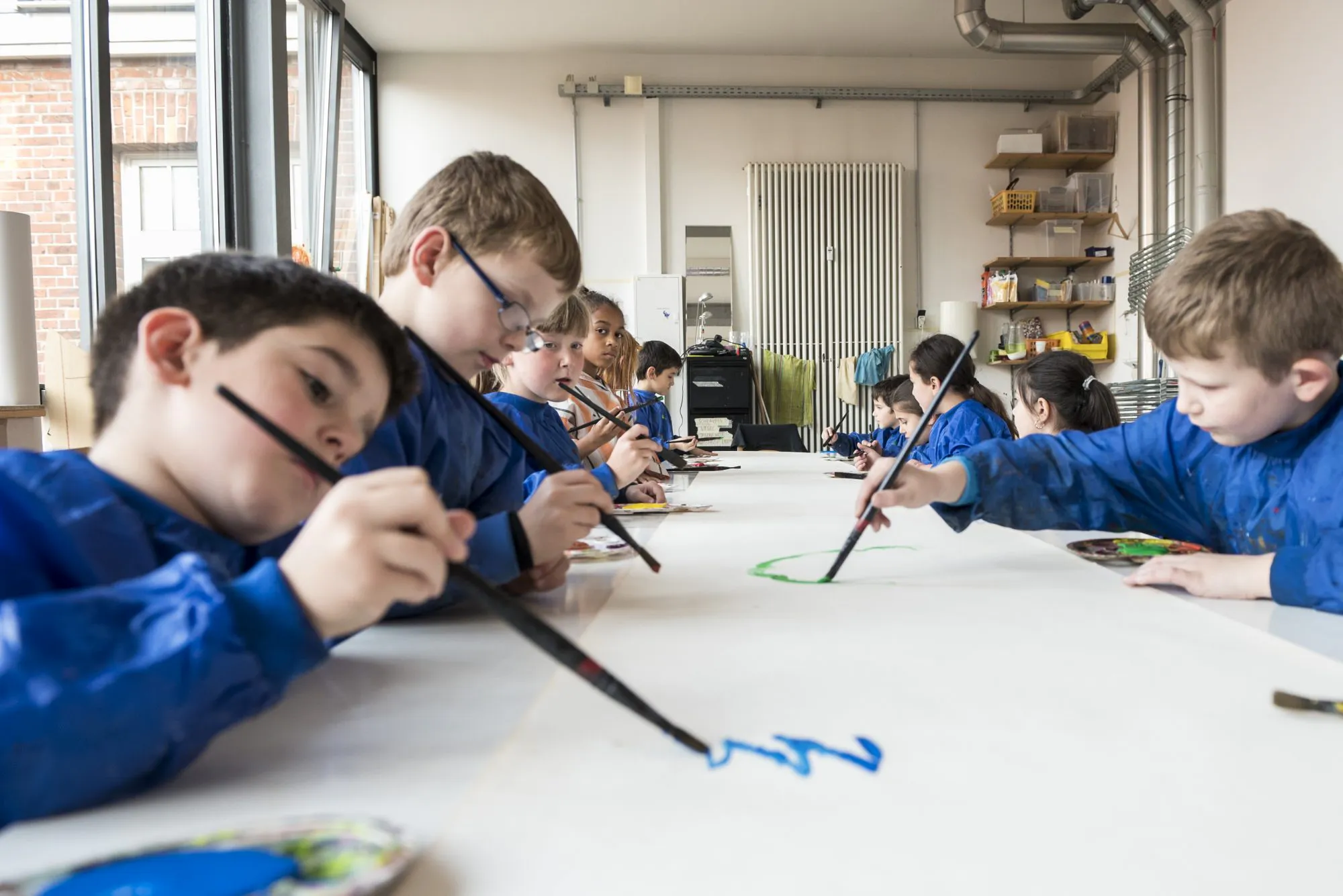 An einem langen Tisch sitzen Kinder in blauen Malkitteln. Vor ihnen liegt großformatiges weißes Papier. Die Kinder halten Pinsel in der Hand und Malen.