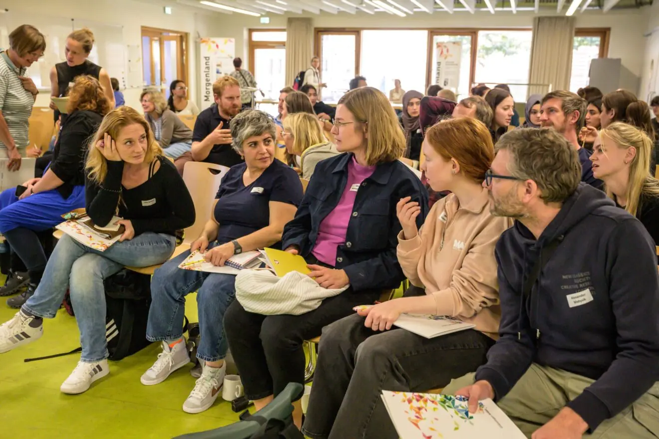 Viele Menschen sitzen in Stuhlreihen bei einer Konferenz. Etliche davon sind in Einzelgespräche vertieft.