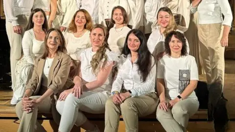 Der Chor Gloria posiert für ein gemeinsames Foto in einem Innenraum mit Holzfußboden und dunkler Wandverkleidung. Alle tragen helle, aufeinander abgestimmte Kleidung in Weiß- und Beigetönen. Einige sitzen in der vorderen Reihe auf einer Holzbank, während die anderen in zwei Reihen dahinter stehen. Die Personen blicken freundlich in die Kamera, das Licht ist warm und gleichmäßig.