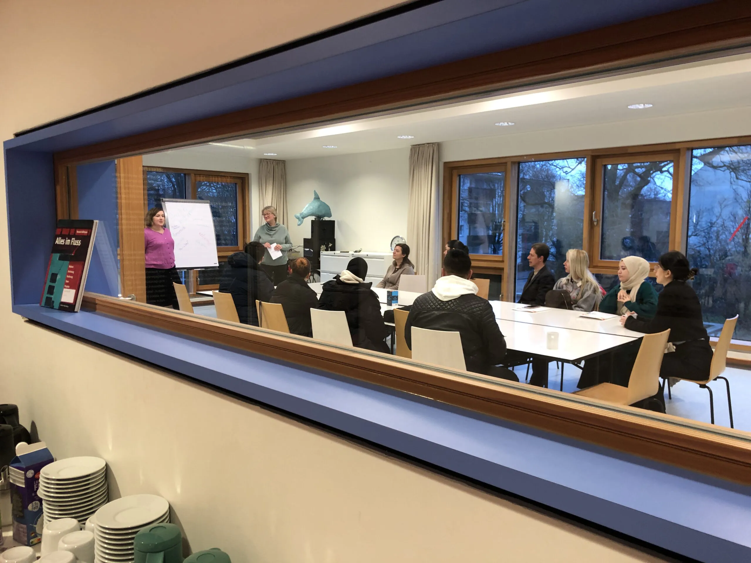 Durch ein Fenster ist eine Gruppe von Personen in einem Raum zu sehen. Zwei Personen stehen vorne und präsentieren an einem Flipchart. Die Teilnehmenden sitzen an einem Tisch und hören aufmerksam zu. Auf der Fensterbank im Vordergrund sind Tassen und Becher sichtbar.
