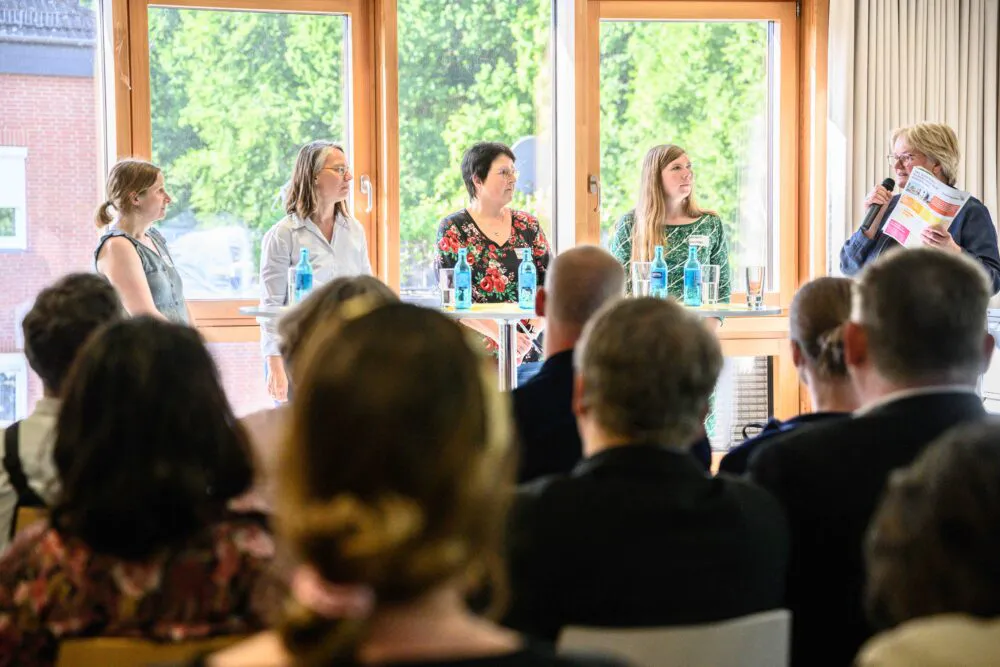 Menschen sitzen in Stuhlreihen, zu sehen sind vor allem die Hinterköpfe. Vor Ihnen befinden sich fünf Personen, die vor einem großen Fester stehen. Vier schauen leicht nach rechts hinüber zu der fünften Person, die in ein Mikrofon spricht.