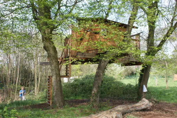 Zwischen mehreren Bäumen befindet sich ein großes Baumhaus. Links daneben steht ein Kind in blauem T-Shirt. 
